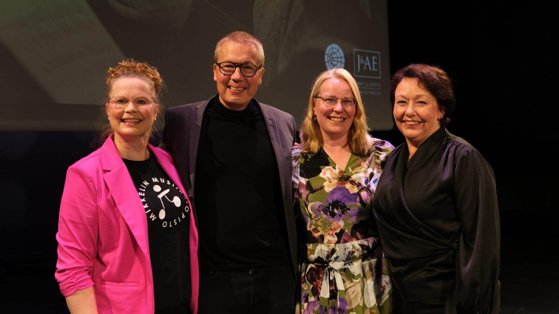 Moderaattori ja kolme panelistia poseeraavat kameralle lavalla.